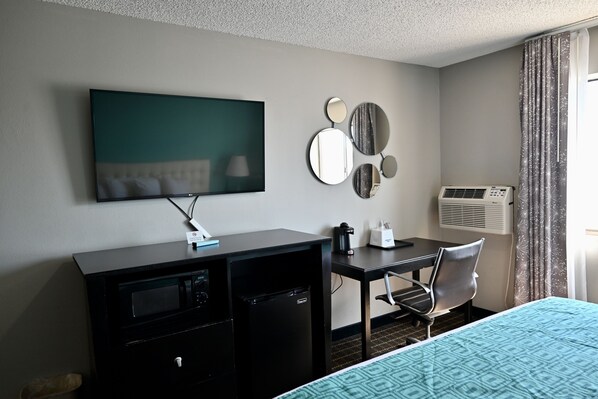 A hotel room with a bed, a desk with a chair, a TV mounted on the wall, a mini-fridge, a coffee maker, and a wall-mounted air conditioner.