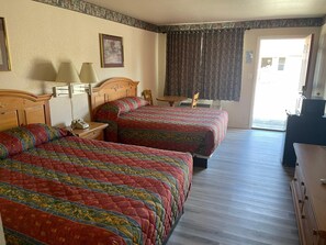 Two double beds, each with a patterned quilt, are set against a wooden headboard. A bedside table with a lamp and a small table with a chair complete the room.