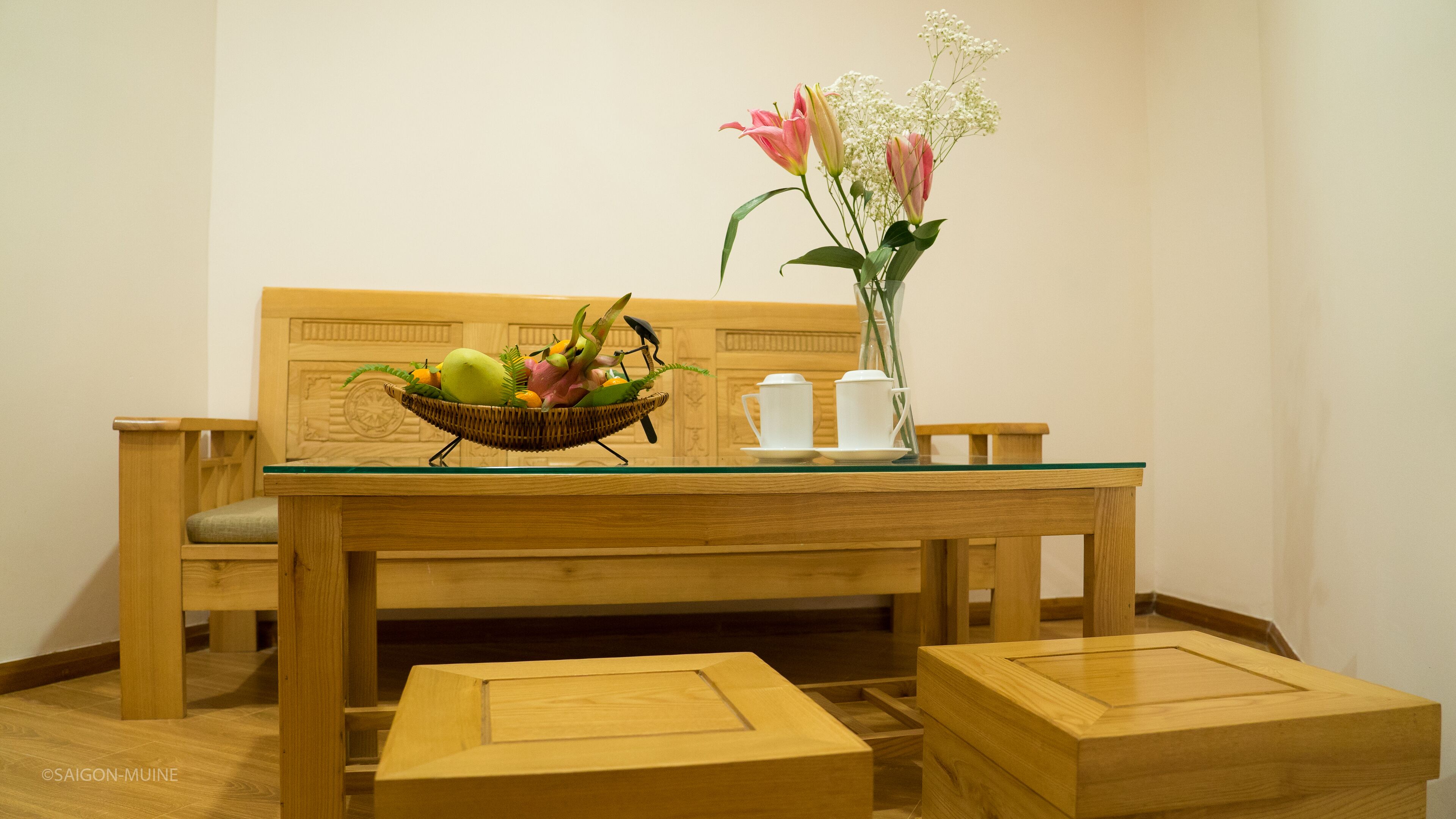 A wooden dining table with a glass top, accompanied by two wooden chairs and a small wooden stool. A wicker basket filled with fruits and vegetables is placed on the table, along with a vase containing flowers and a set of white cups and saucers.