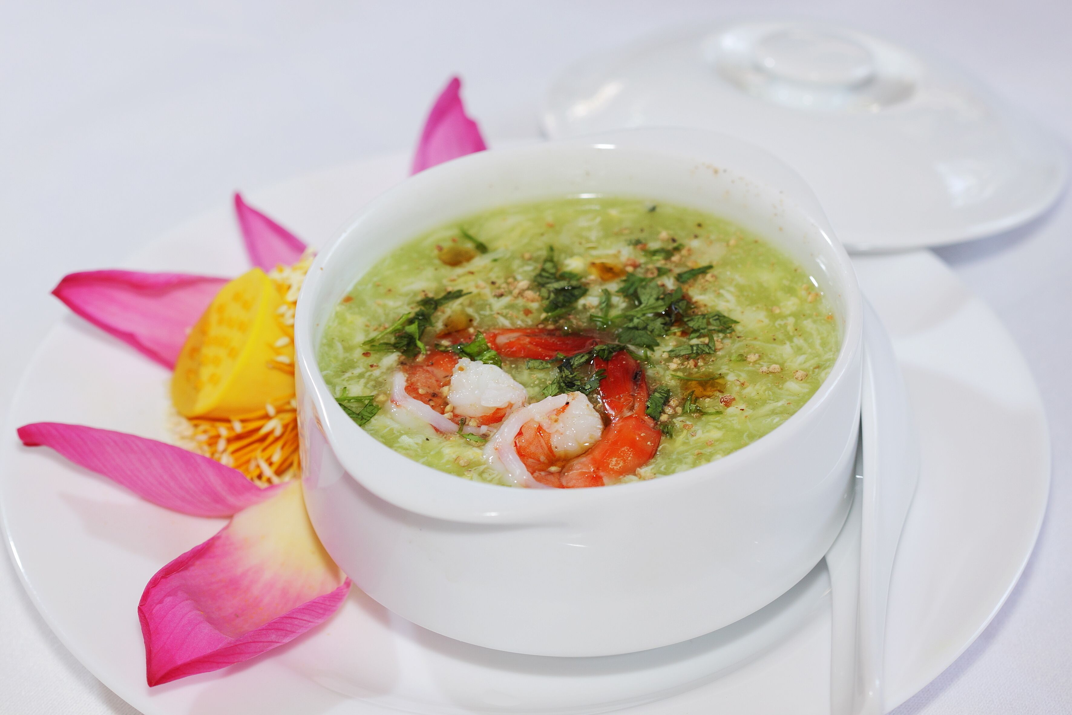 Un bol de soupe aux crevettes et aux herbes, servi dans une assiette blanche, agrémenté d’un pétale de fleur rose et d’un fruit jaune.