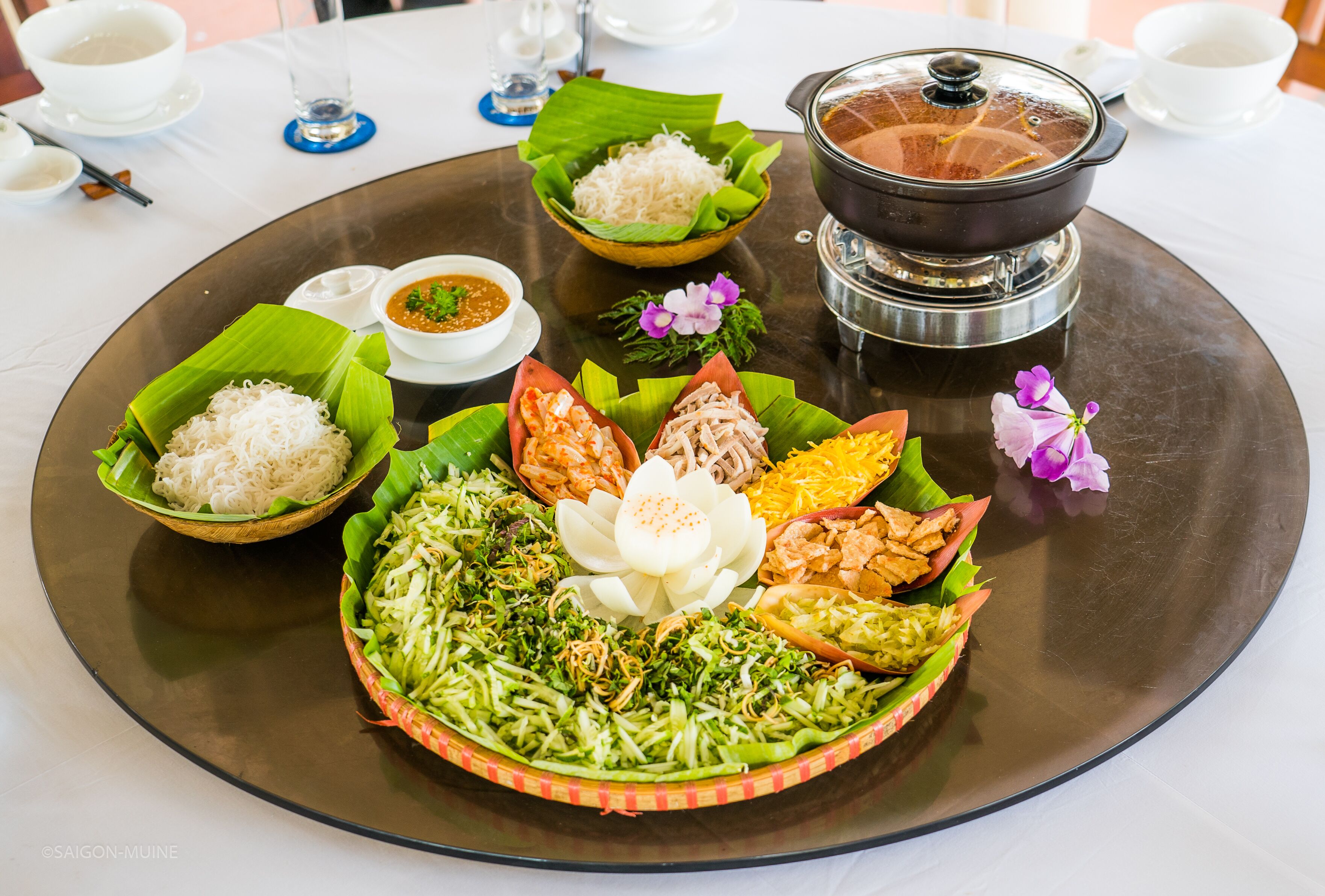 A traditional meal setup with various dishes on a large platter, including rice, vegetables, and meat, accompanied by a pot of soup and small bowls of sauces.