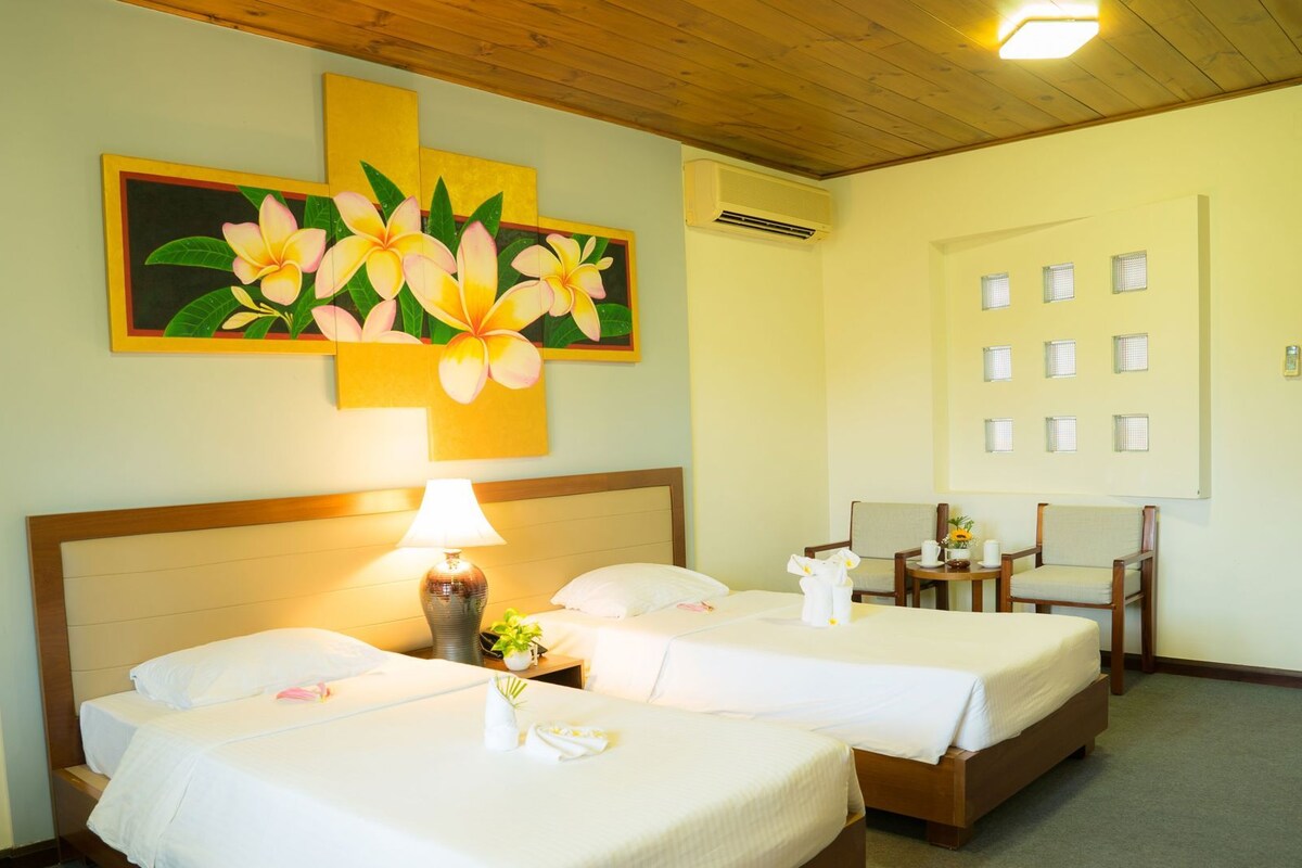 A hotel room with two beds, a nightstand with a lamp, a painting of flowers, and a wall with multiple small windows.