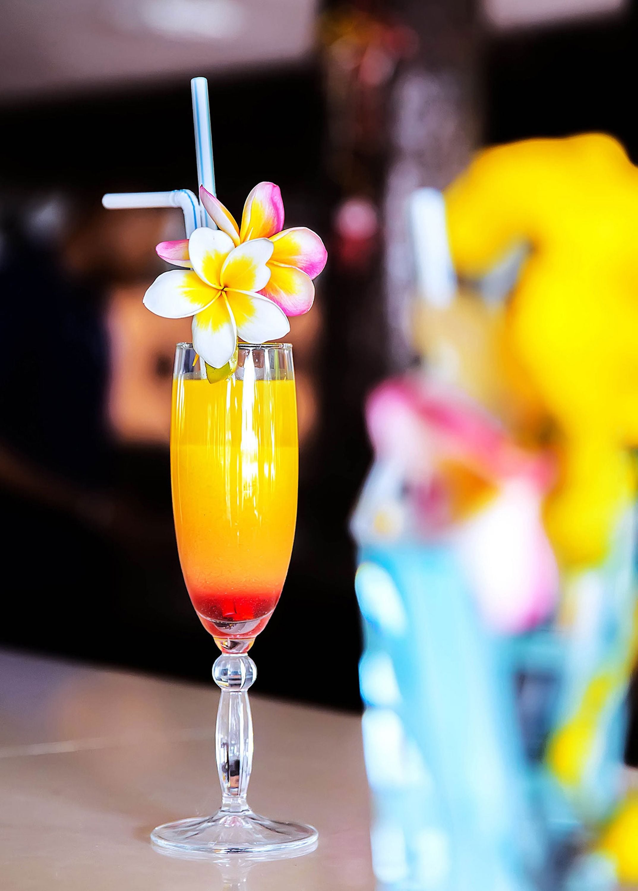 Ein Cocktailglas mit einem schichtförmigen Getränk, einem Strohhalm und einer Blütenblumengarnitur auf einer Bar.