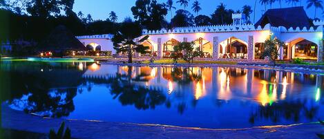 A resort with a large reflective pool, a thatched-roof building, and a white-walled structure with multiple rooms.