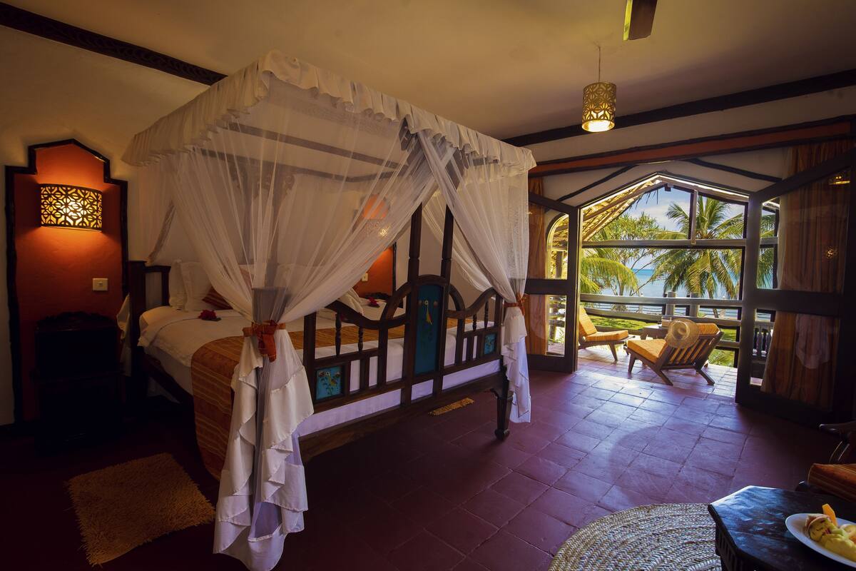 A bedroom with a four-poster bed, a nightstand, a bedside lamp, and a view of palm trees through a large window.