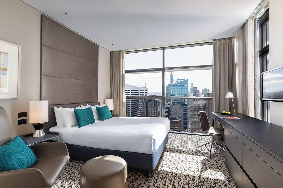 A modern hotel room with a large bed, a desk, a chair, and a view of the cityscape.