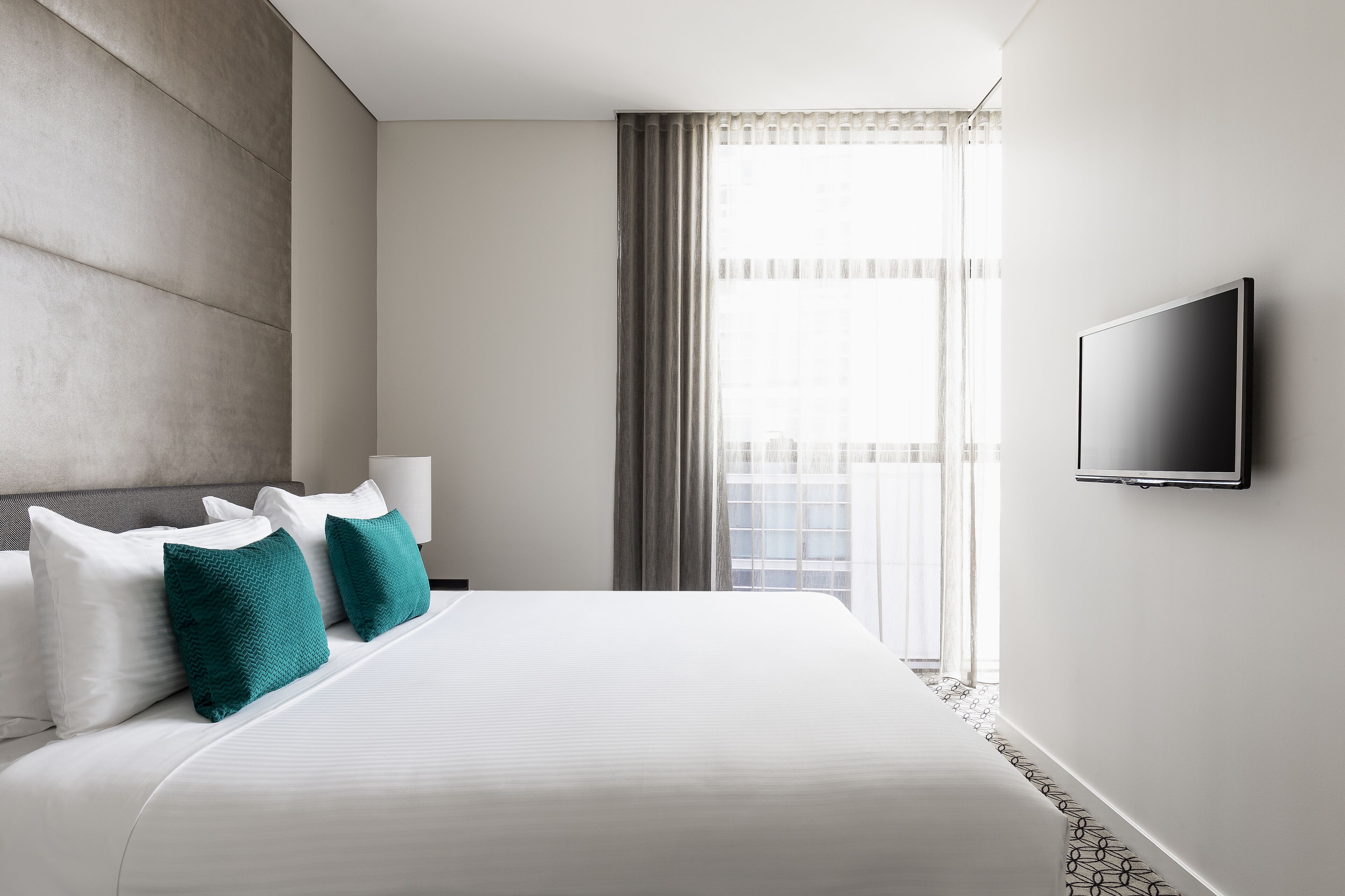 A hotel room with a large bed, a flat-screen TV mounted on the wall, and a window with curtains.