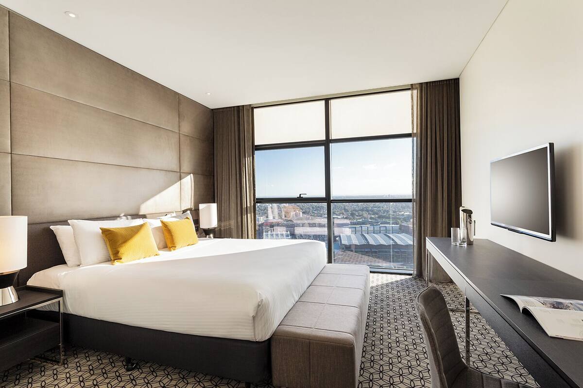 A modern hotel room with a large bed, a TV, a desk, and a view of the city.