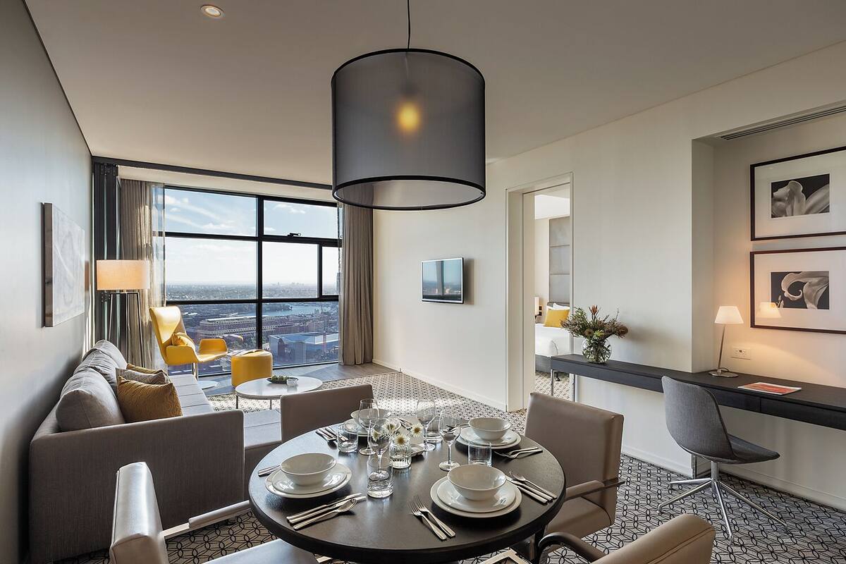 A modern hotel room with a dining area, a sofa set, a large window with a city view, and a desk with a lamp.
