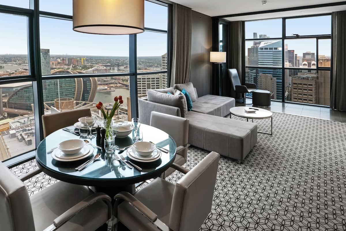 A modern hotel room with a panoramic city view, a dining area with a round table, and a cozy seating area with a sofa and armchairs.
