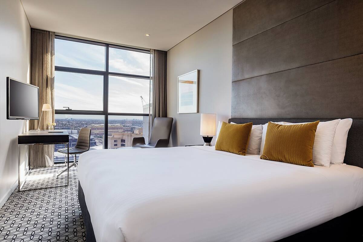 A hotel room with a large bed, a desk, a chair, a TV, and a view of the city.