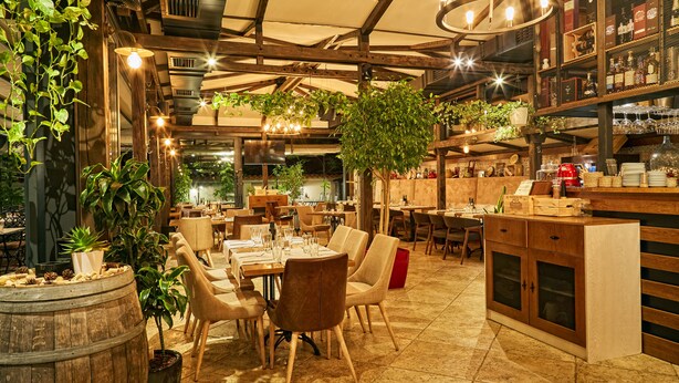 Un restaurant avec des poutres en bois, des plantes suspendues et un bar doté d’étagères remplies de bouteilles.