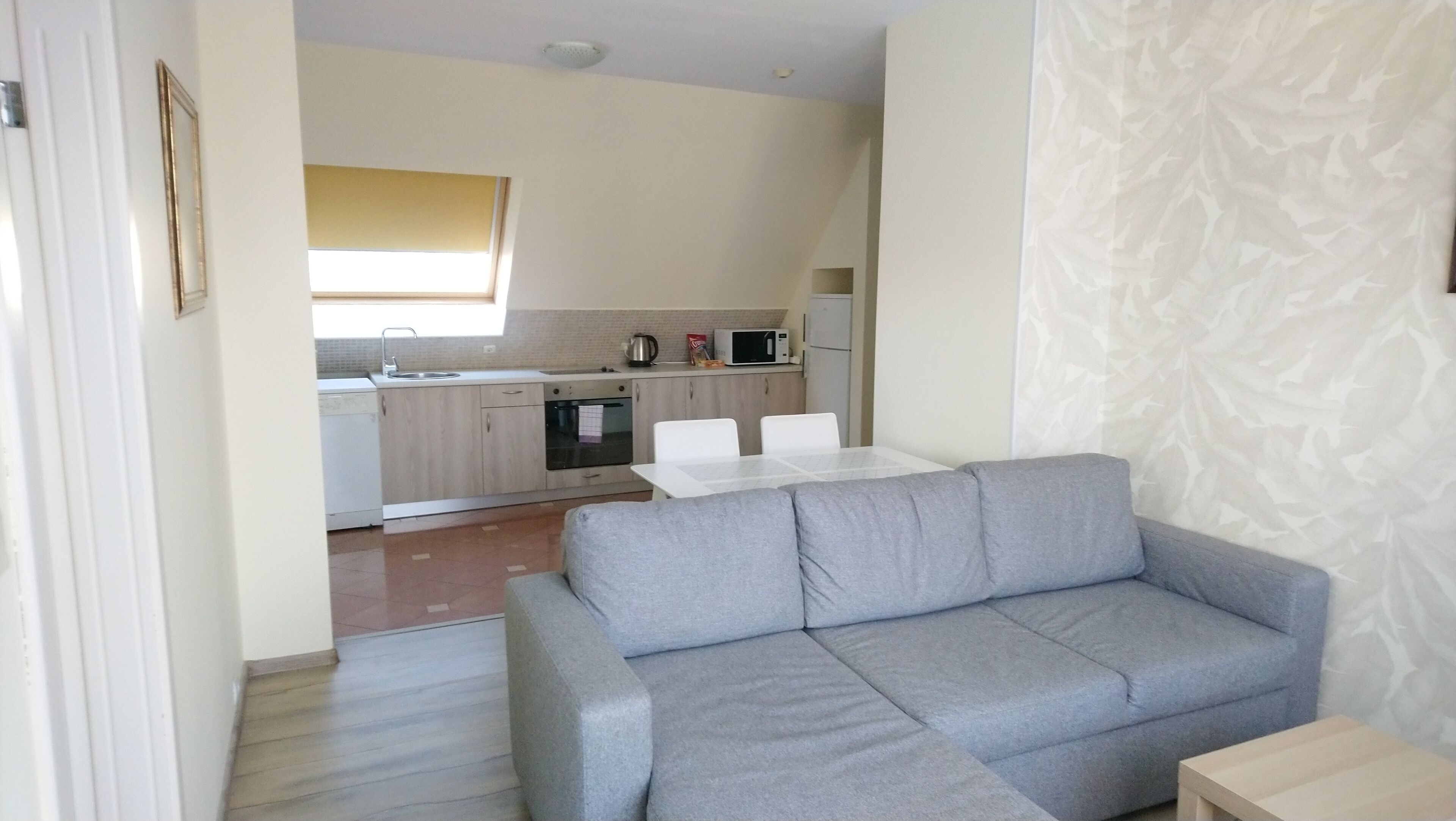 A modern living room with a grey sofa and a dining table, a kitchen with light wood cabinets, a microwave, and a kettle.