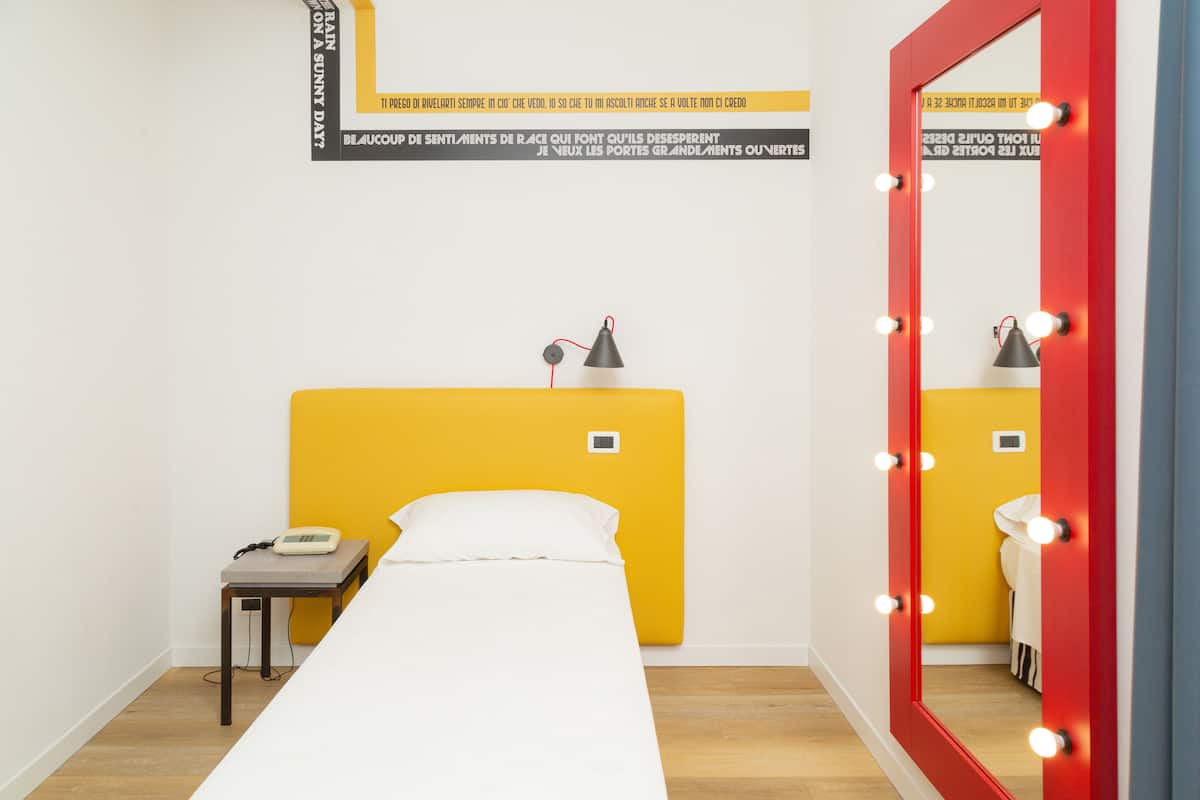 A room with a bed, a small table with a lamp, a mirror with lights, and a wall with text.