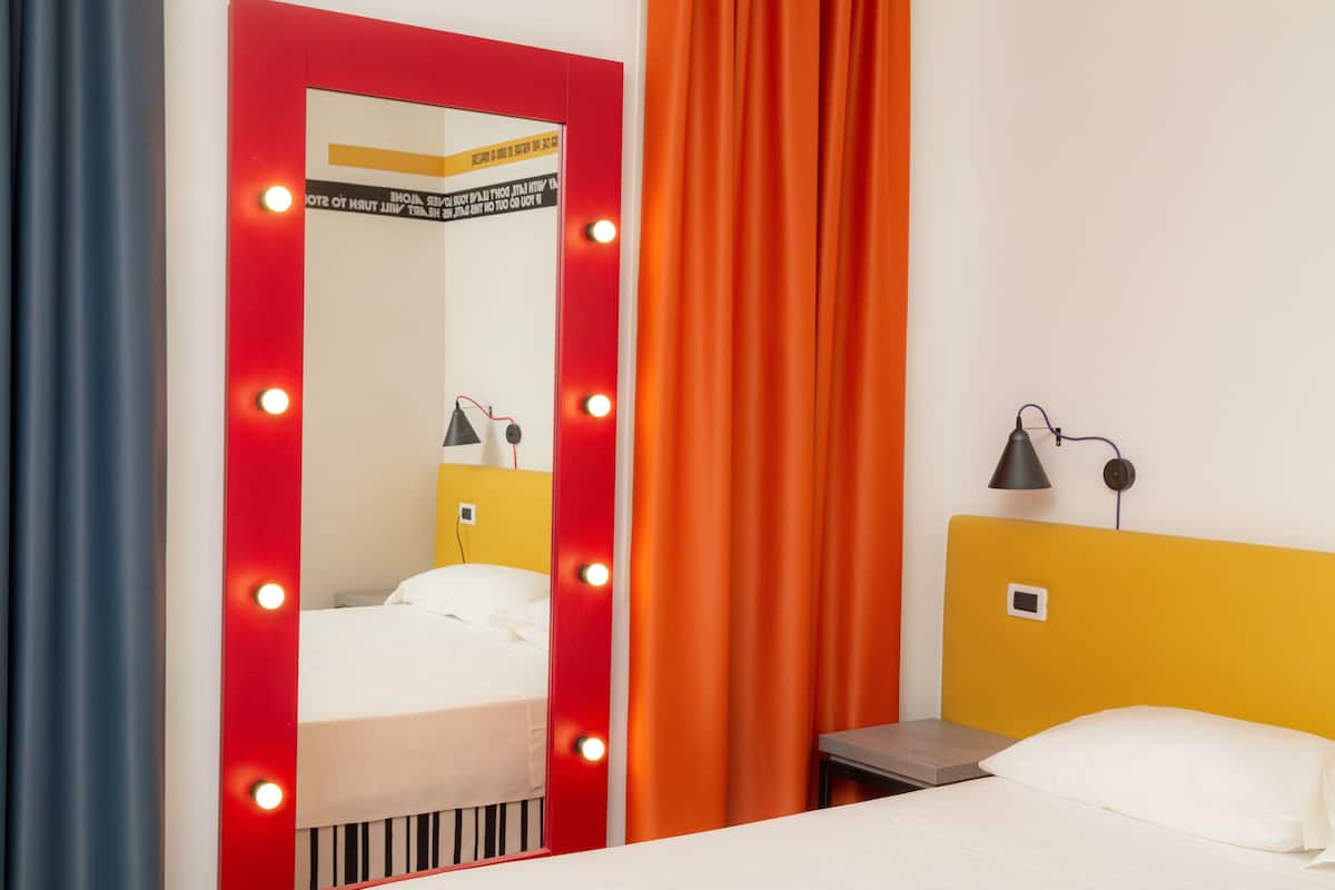 A hotel room with a bed, a mirror with lights, a nightstand, and a lamp.