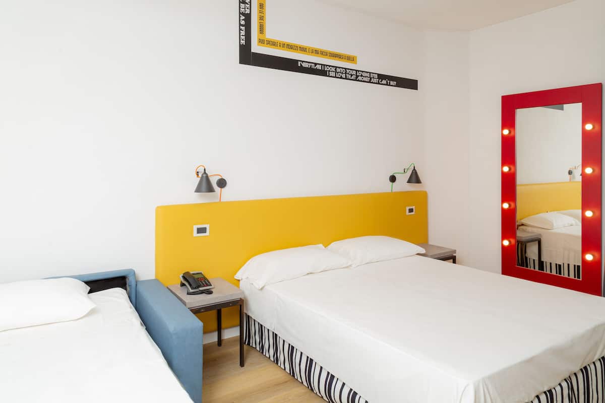 A hotel room with a yellow headboard, a bed with white bedding, a bedside table with a lamp, a mirror with lights, and a small desk with a telephone.