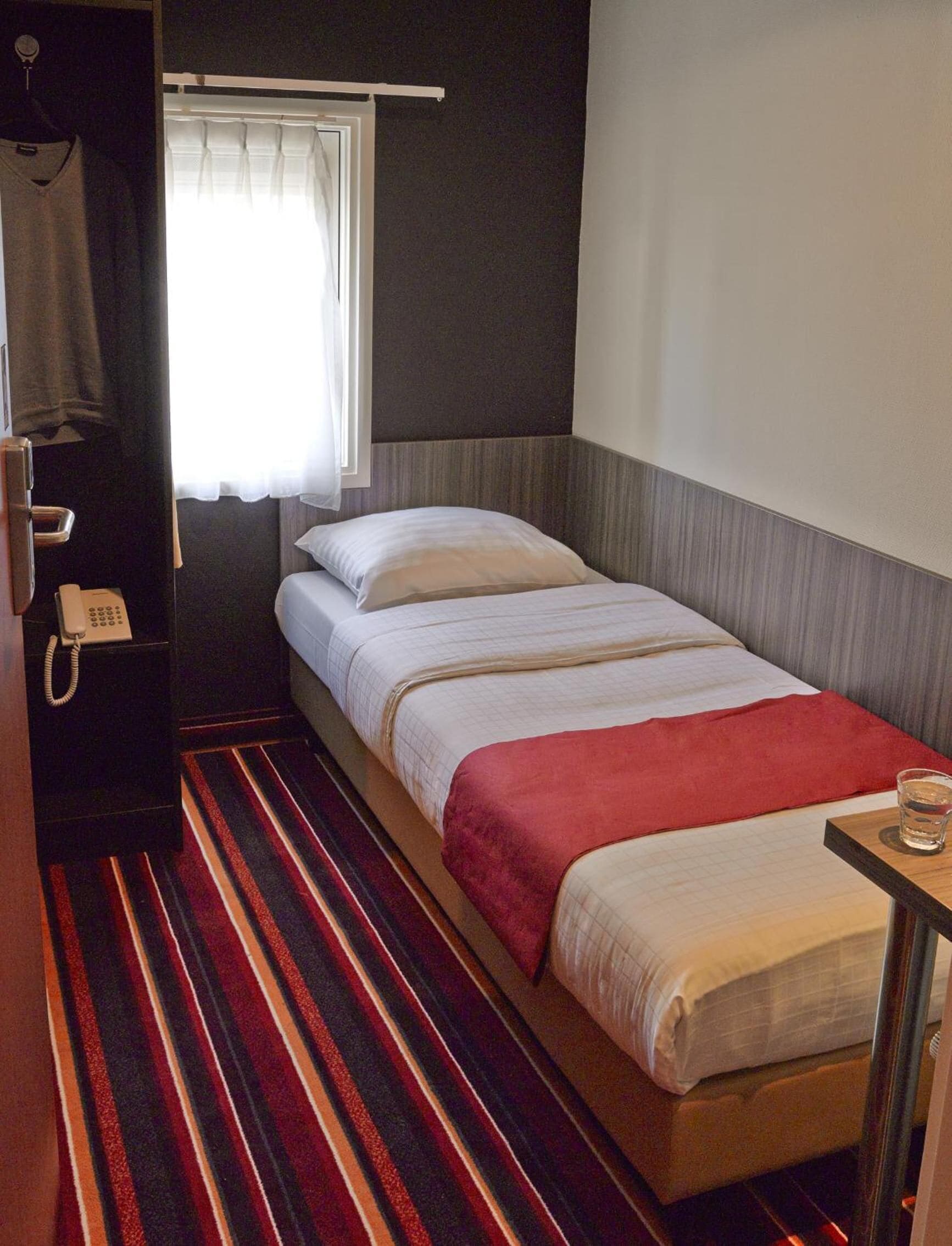Una cama individual con alfombra a rayas, una mesita de noche con un vaso y un teléfono a la izquierda.
