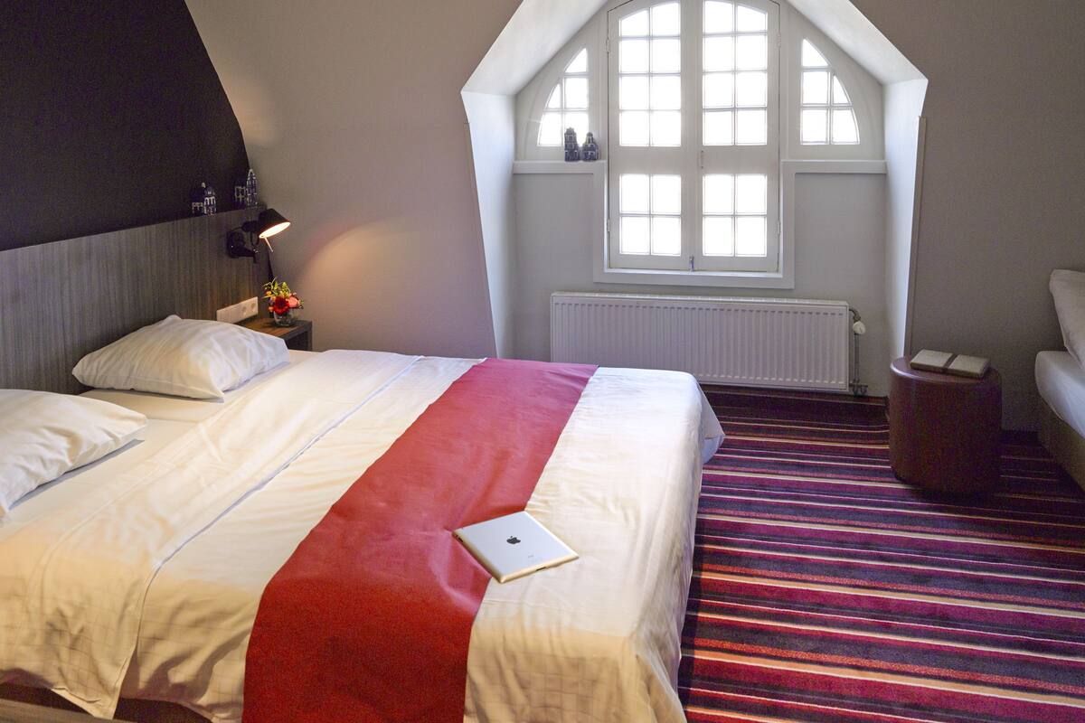 Quarto de hotel com uma cama grande, um tapete listrado em vermelho e branco, uma janela com persianas brancas e uma pequena mesa com abajur.