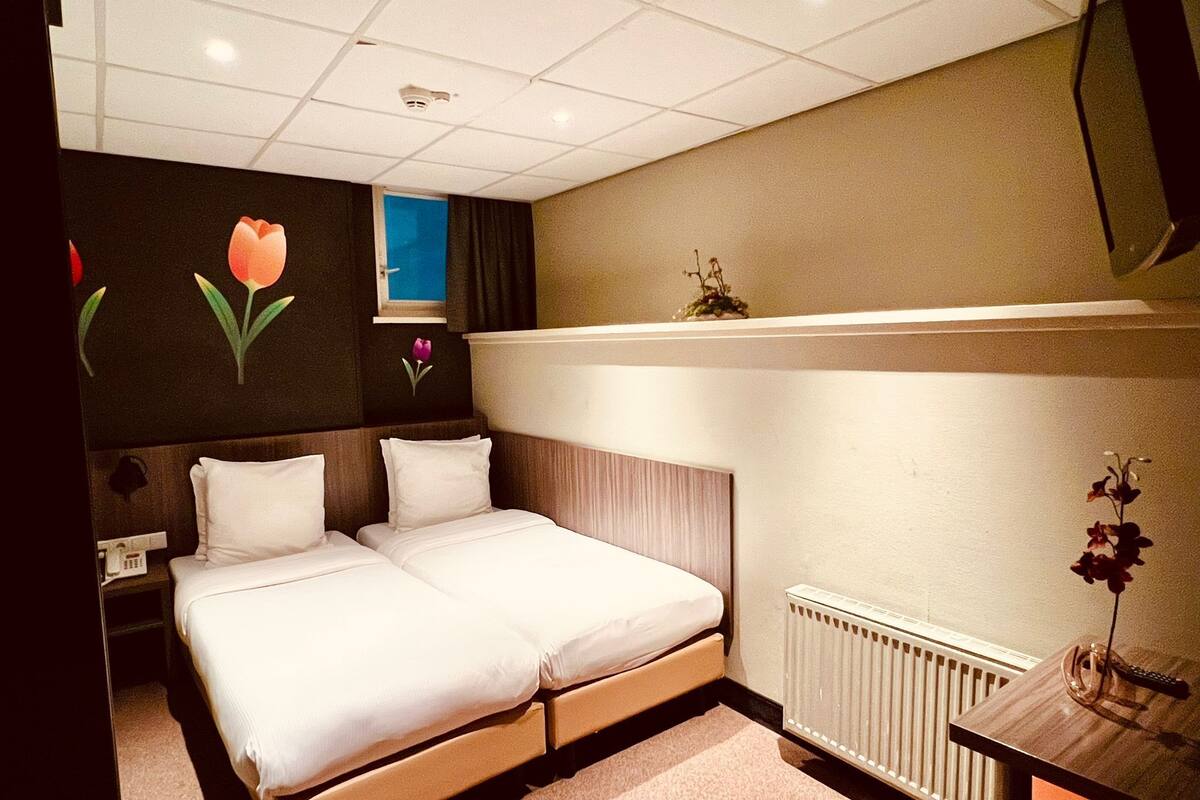 Quarto de hotel com uma cama de casal, uma pequena mesa de cabeceira, um radiador e uma televisão fixada na parede.