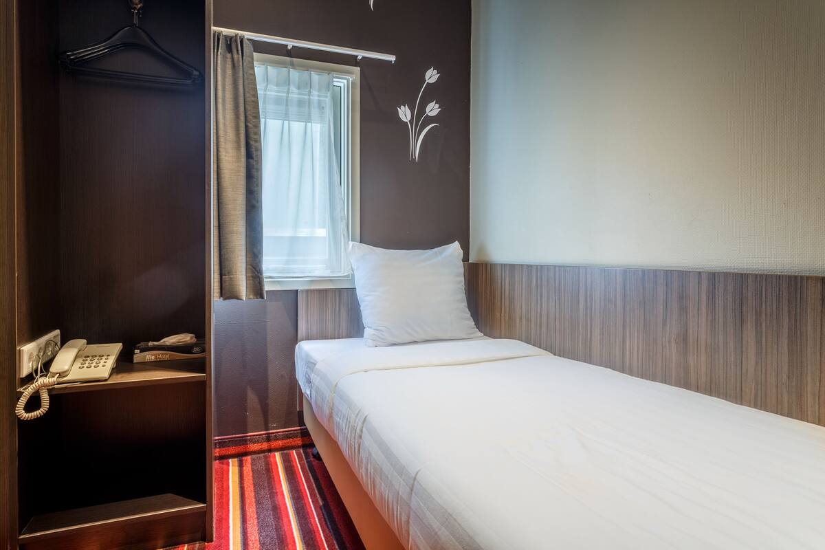 Quarto de hotel com cama, telefone, janela com cortinas e luminária fixada na parede.