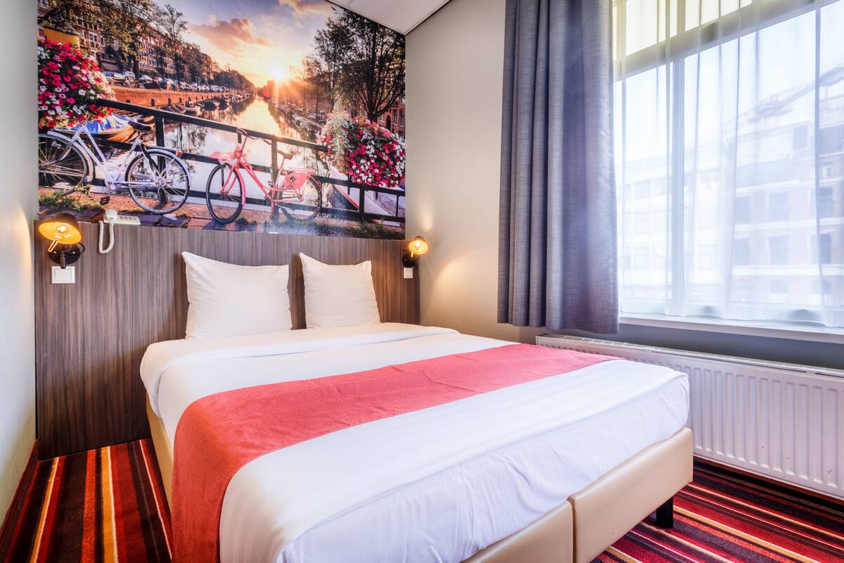 Quarto de hotel com uma cama grande, um mural da cidade na parede, uma janela com cortinas e um tapete listrado.