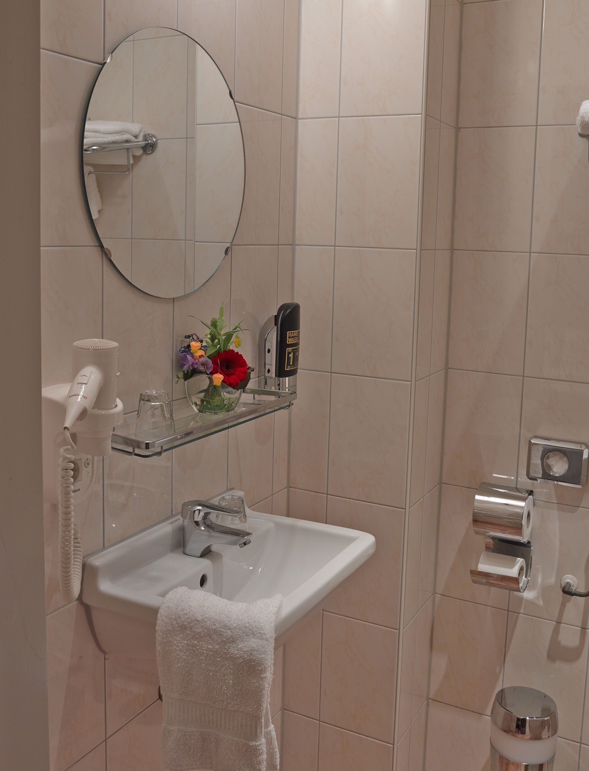 A bathroom with a white sink, a mirror, a hand dryer, and a glass shelf with a vase of flowers.