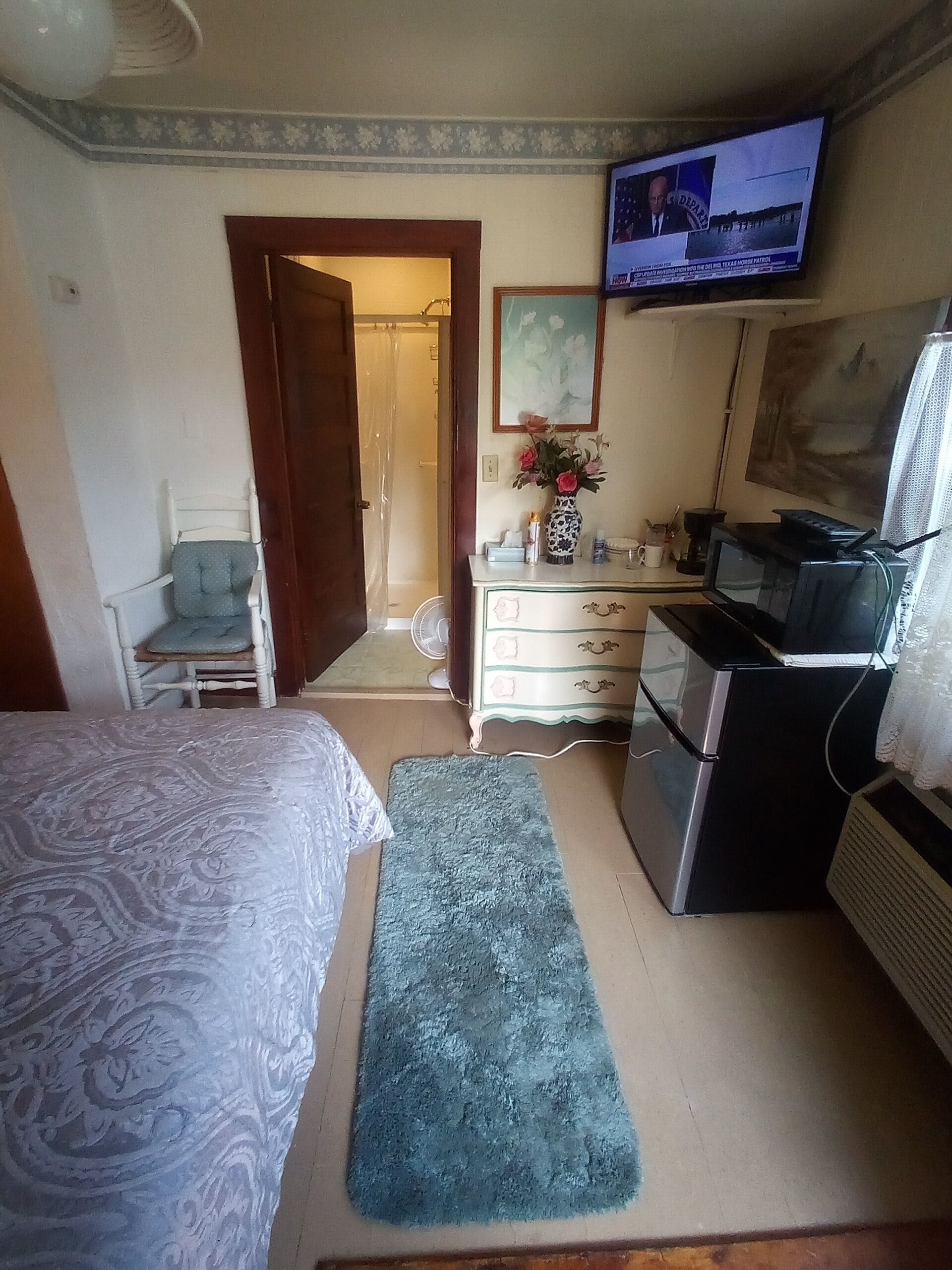 A bedroom with a bed, a desk with a computer, a TV, and a bathroom visible through an open door.
