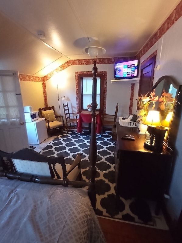 A compact hotel room with a bed, a desk with a lamp, a television, and a small kitchenette.