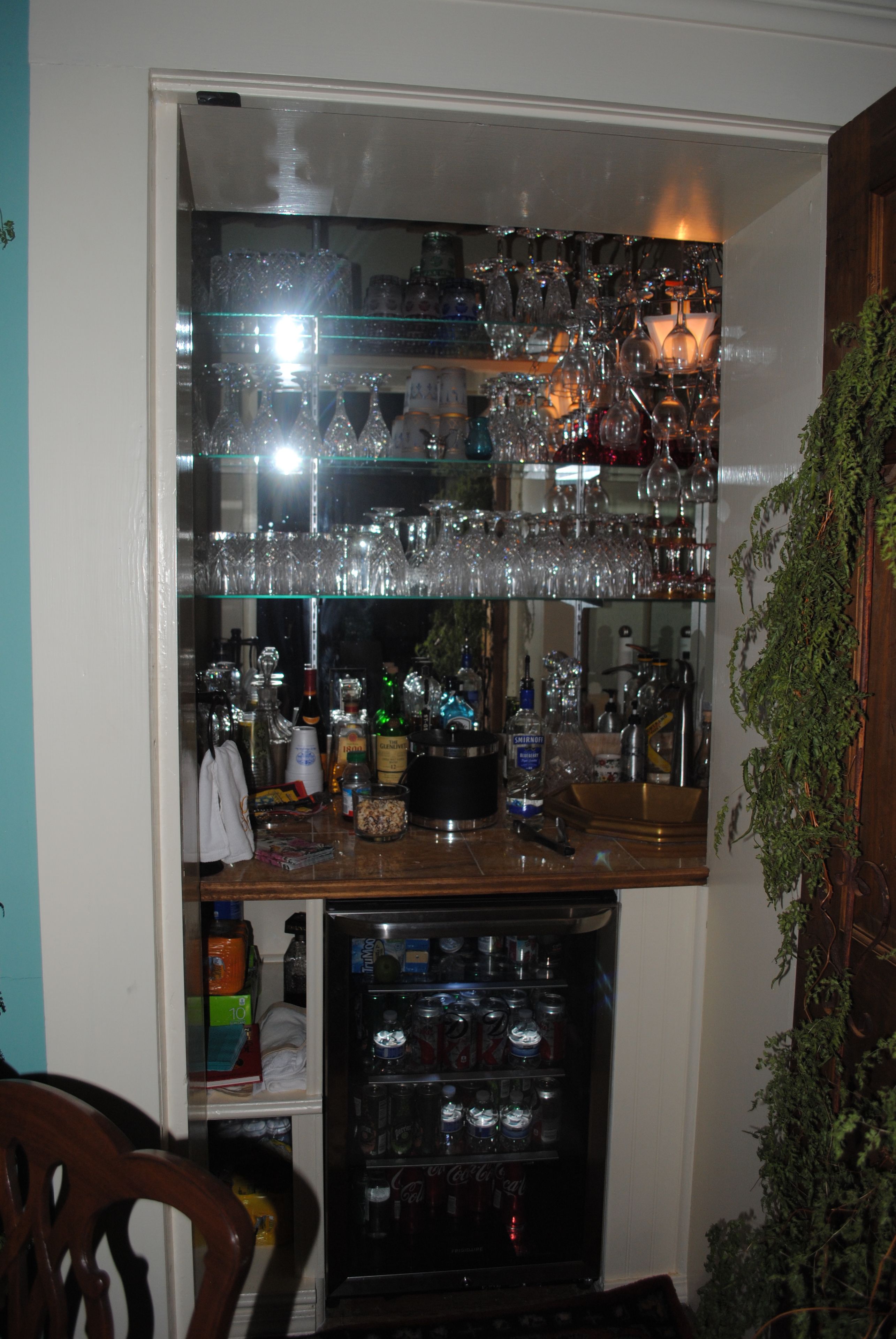 A bar area with shelves of glassware and bottles, a refrigerator with beverages, and a wooden chair.