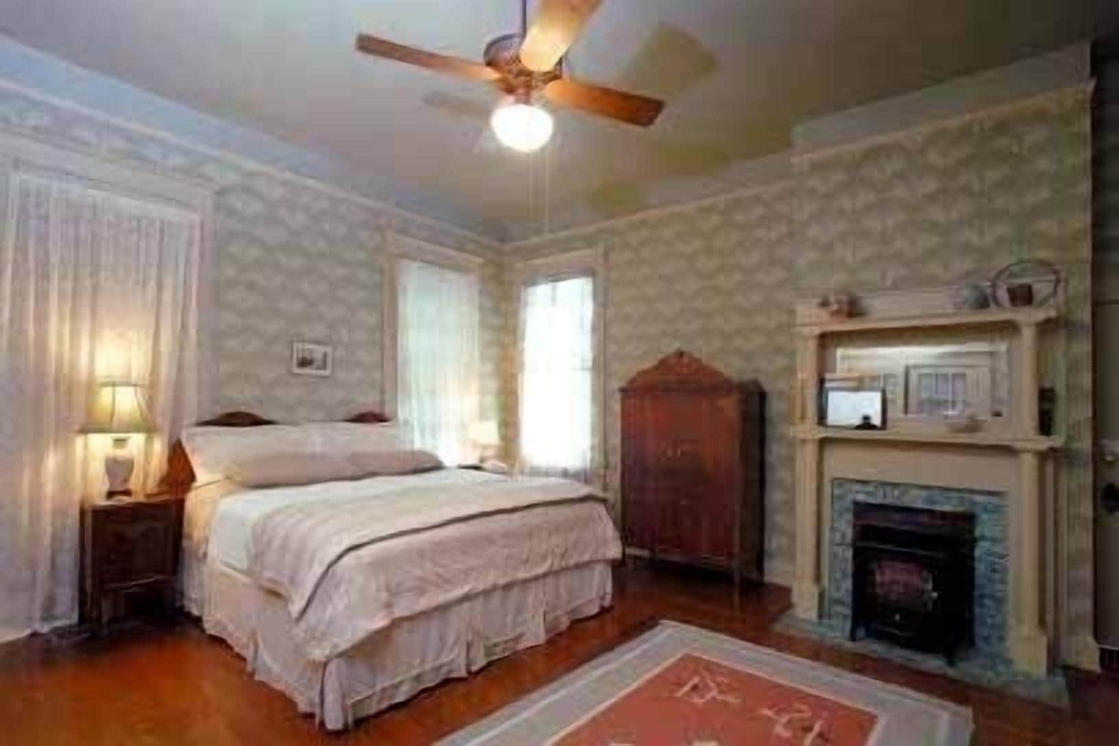 A bedroom with a bed, a nightstand, a wooden wardrobe, a fireplace, and a ceiling fan.