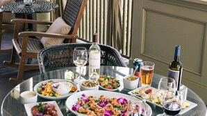 A glass table set with various dishes, wine glasses, and a bottle of wine, surrounded by wicker chairs and a window with curtains. - Princess Anne Boutique Hotel (Asheville)