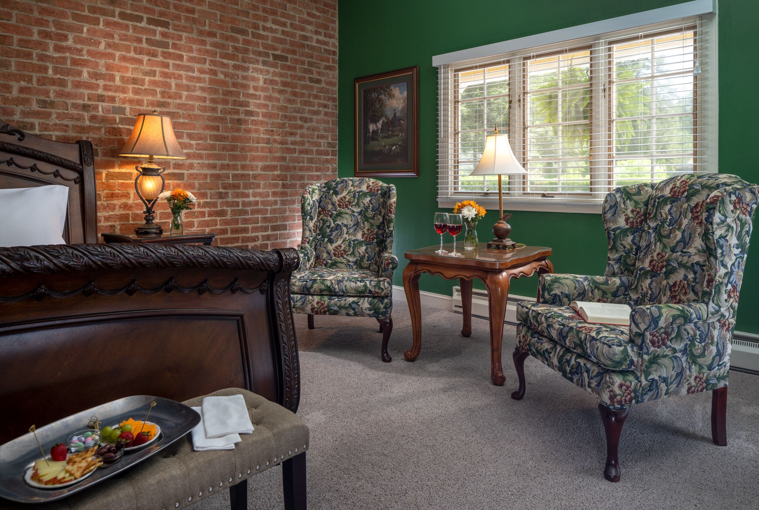 A room with a brick wall, green walls, a bed with a wooden headboard, two floral armchairs, a small wooden table with a lamp, and a window with blinds.