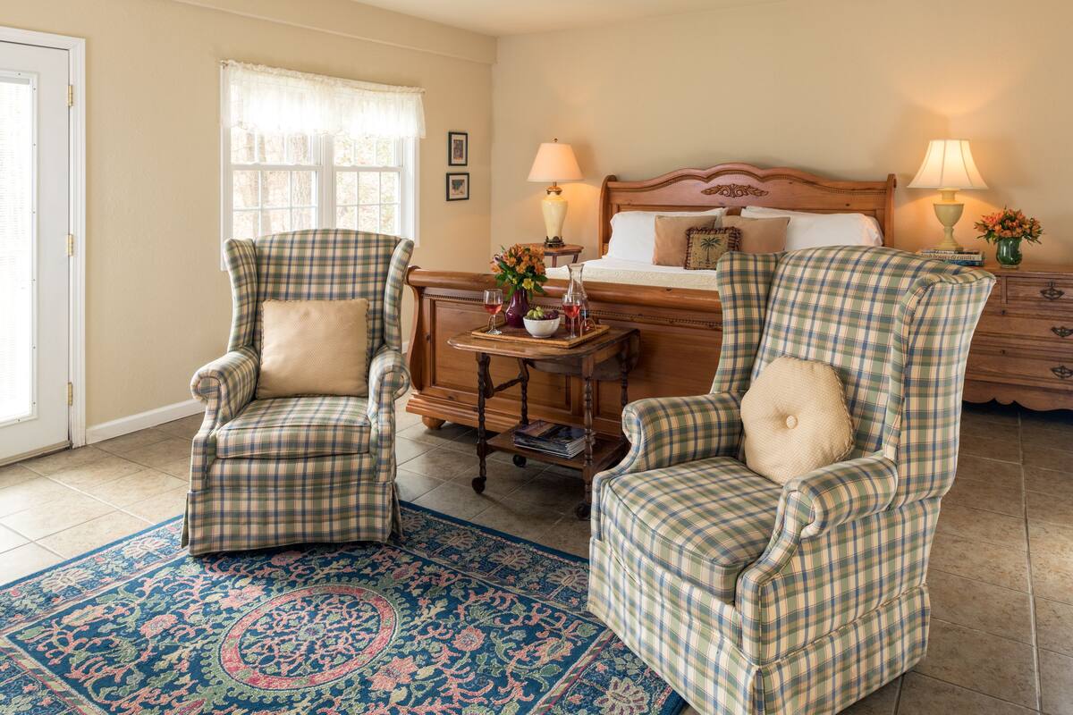 A bedroom with two plaid armchairs, a wooden nightstand with lamps, a bed with a wooden headboard, and a patterned rug.