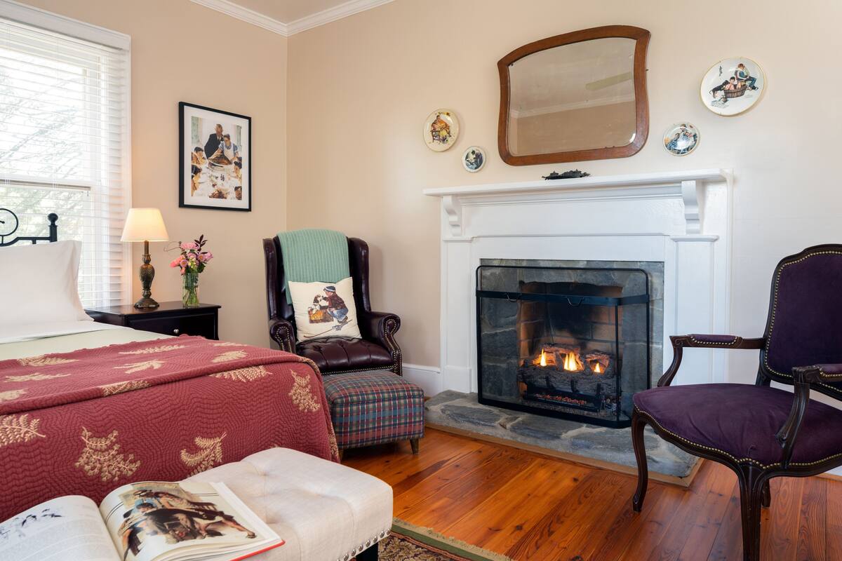 A cozy room with a fireplace, a bed, a chair, and a table with a lamp and flowers.