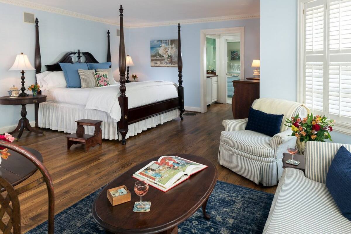 A bedroom with a four-poster bed, a sitting area with a striped armchair, and a coffee table with a book and glasses.