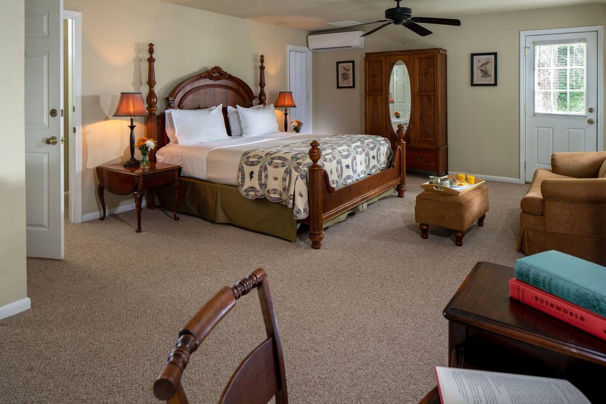 A bedroom with a wooden bed, two bedside tables with lamps, a large mirror, a wooden wardrobe, a chair, a small table with books, and a door with a window.