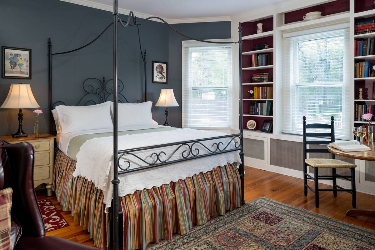 A four-poster bed with a white canopy, a wooden chair, a small table with a book, and a bookshelf filled with books.