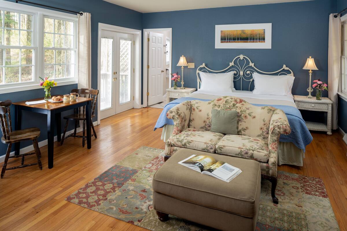 A bedroom with a bed, a floral sofa, a wooden desk with chairs, and a window with curtains.