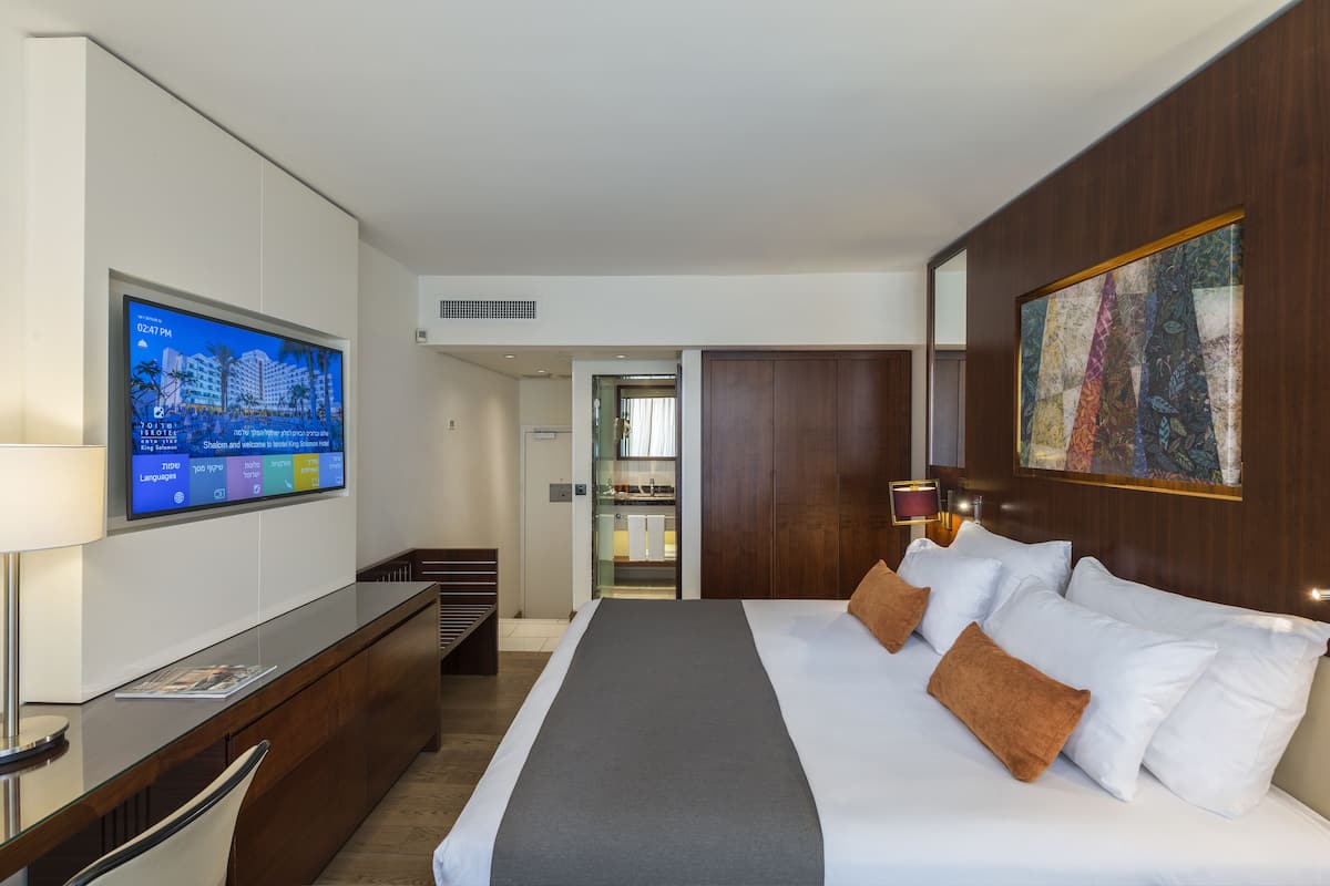 Quarto de hotel moderno com uma cama grande, uma TV fixada na parede, um guarda-roupa de madeira e uma porta de vidro para o banheiro.