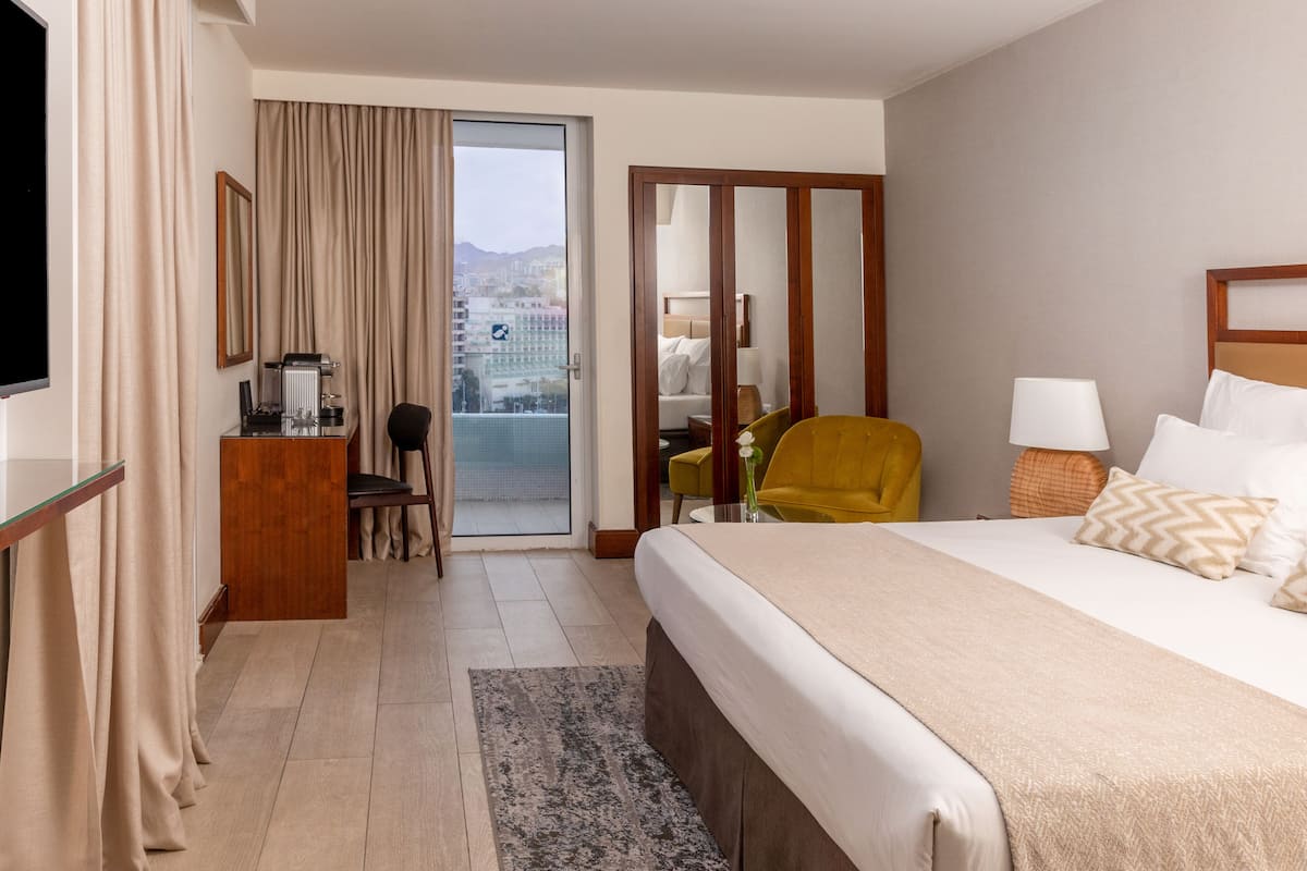 Quarto de hotel com cama, escrivaninha, cadeira, televisão, janela com cortinas e vista para a cidade.