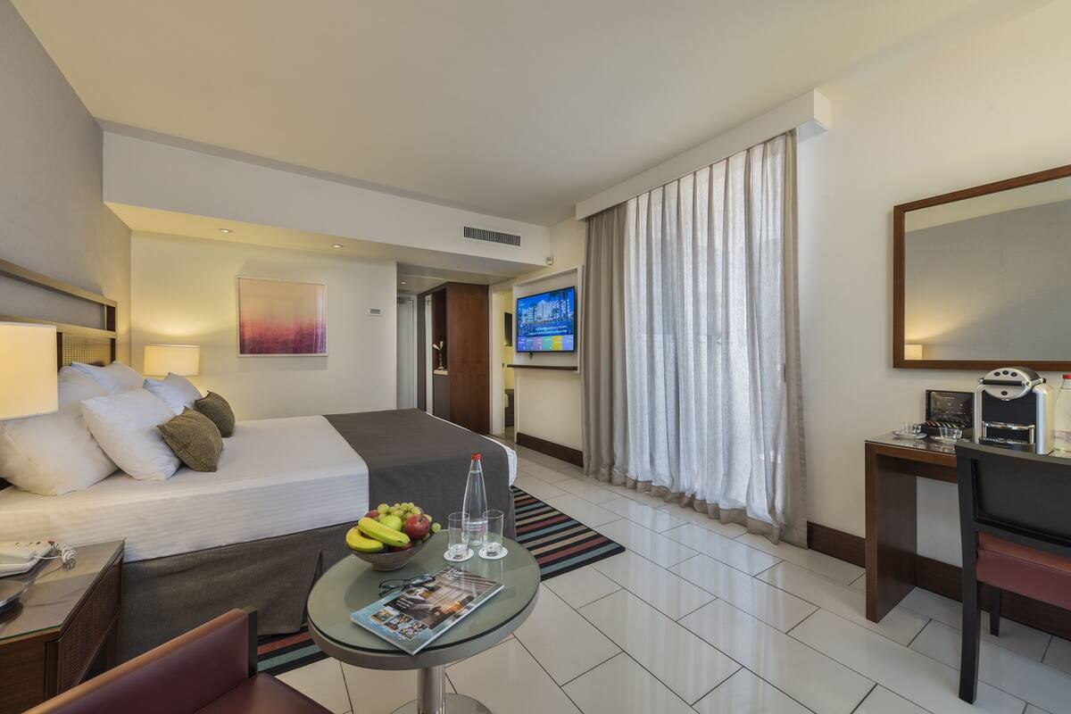 Quarto de hotel com cama, secretária com máquina de café, televisão e uma tigela de fruta na mesa.