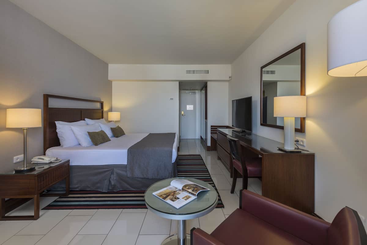 Quarto de hotel com cama, mesas de cabeceira, escrivaninha, cadeira, televisão e espelho.