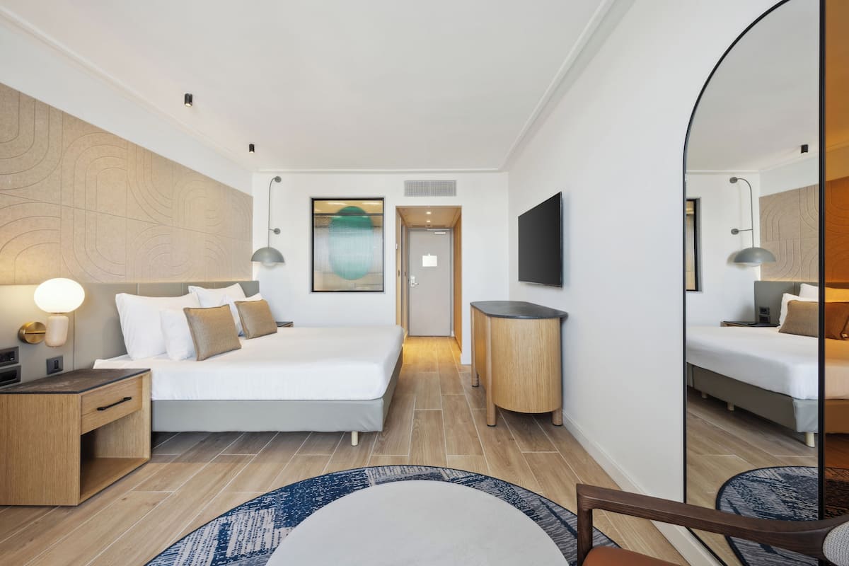 Quarto de hotel moderno com uma cama grande, mesa de cabeceira, espelho e televisão de tela plana.