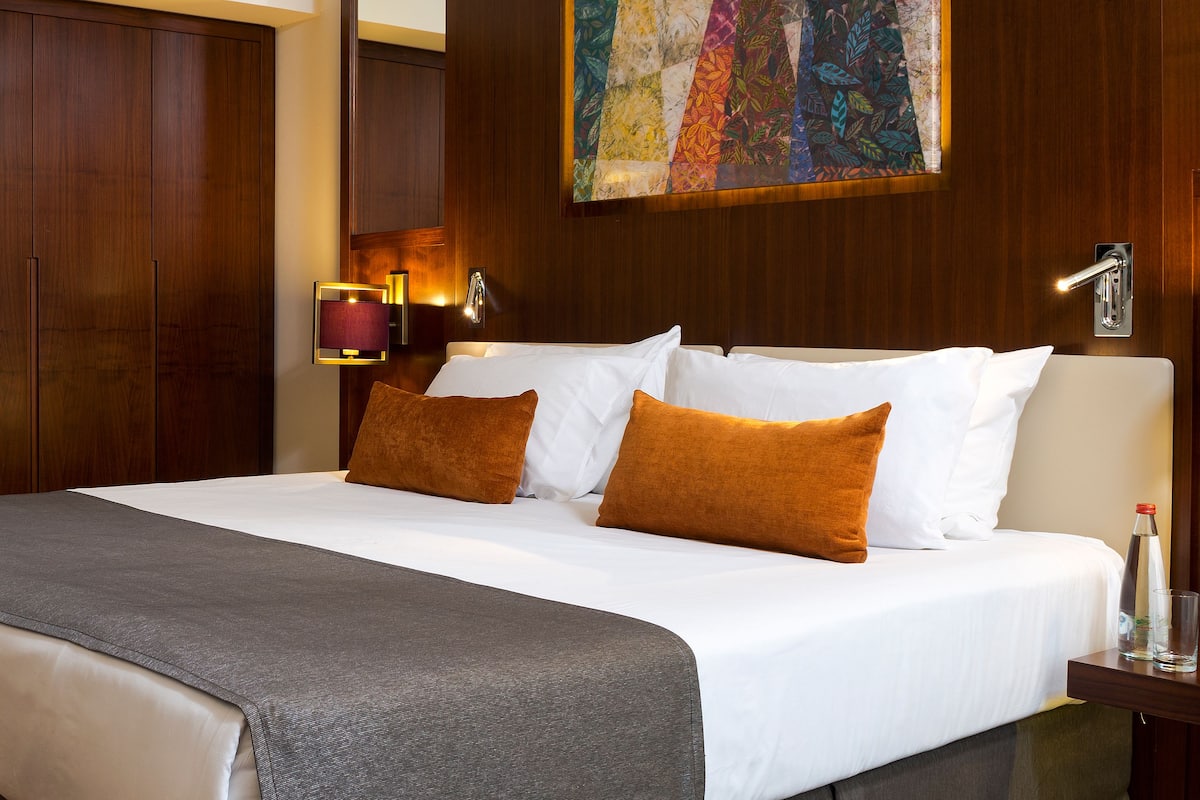 Cama bem arrumada com almofadas brancas e castanhas, um cobertor cinza e cabeceira de madeira. Há uma luminária fixada na parede e um quadro sobre a cama.