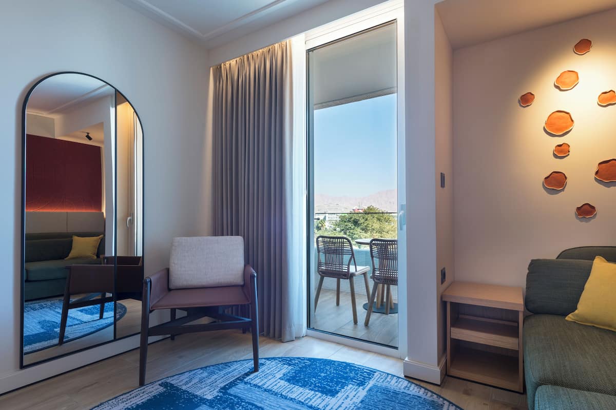 Quarto de hotel moderno com uma grande porta de vidro deslizante para uma varanda, uma área de estar com sofá e cadeiras, e uma parede decorativa com padrões circulares.
