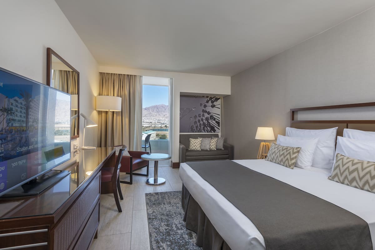 Quarto de hotel com cama, escrivaninha, televisão, cadeira, mesinha e vista para as montanhas.
