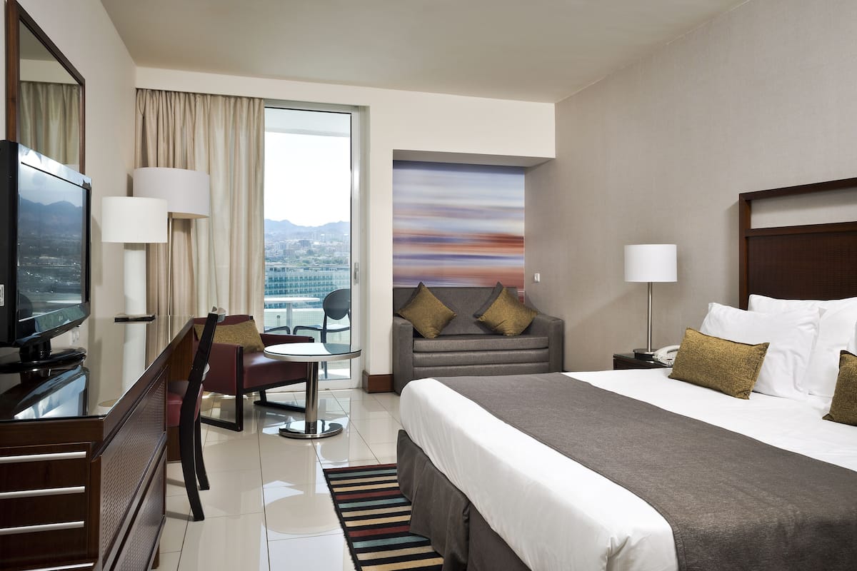 Quarto de hotel com cama, escrivaninha com cadeira, televisão e vista para a cidade pela janela.