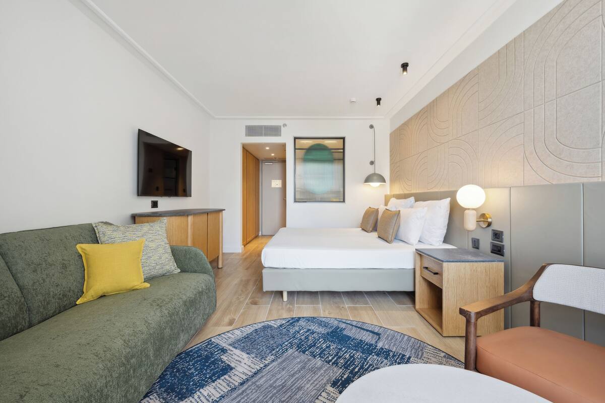Quarto de hotel moderno com sofá verde, cama com roupa de cama branca, mesa de cabeceira de madeira e uma pequena escrivaninha.