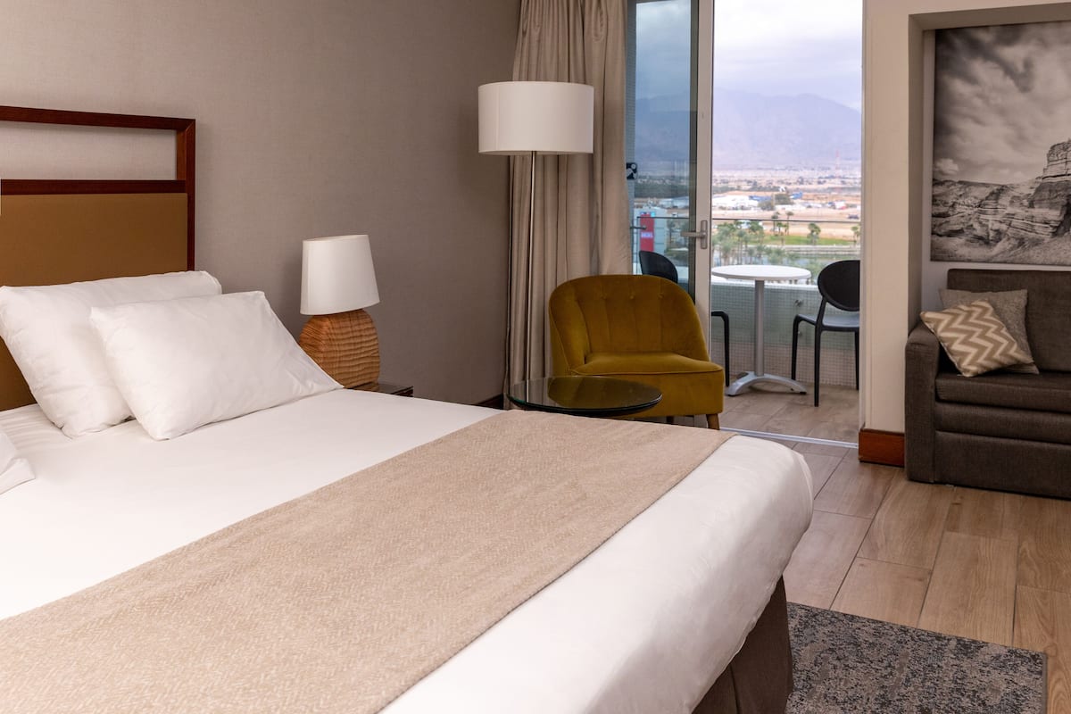 Quarto de hotel com uma cama grande, um sofá, uma poltrona, uma mesa e vista para a cidade e montanhas pela janela.