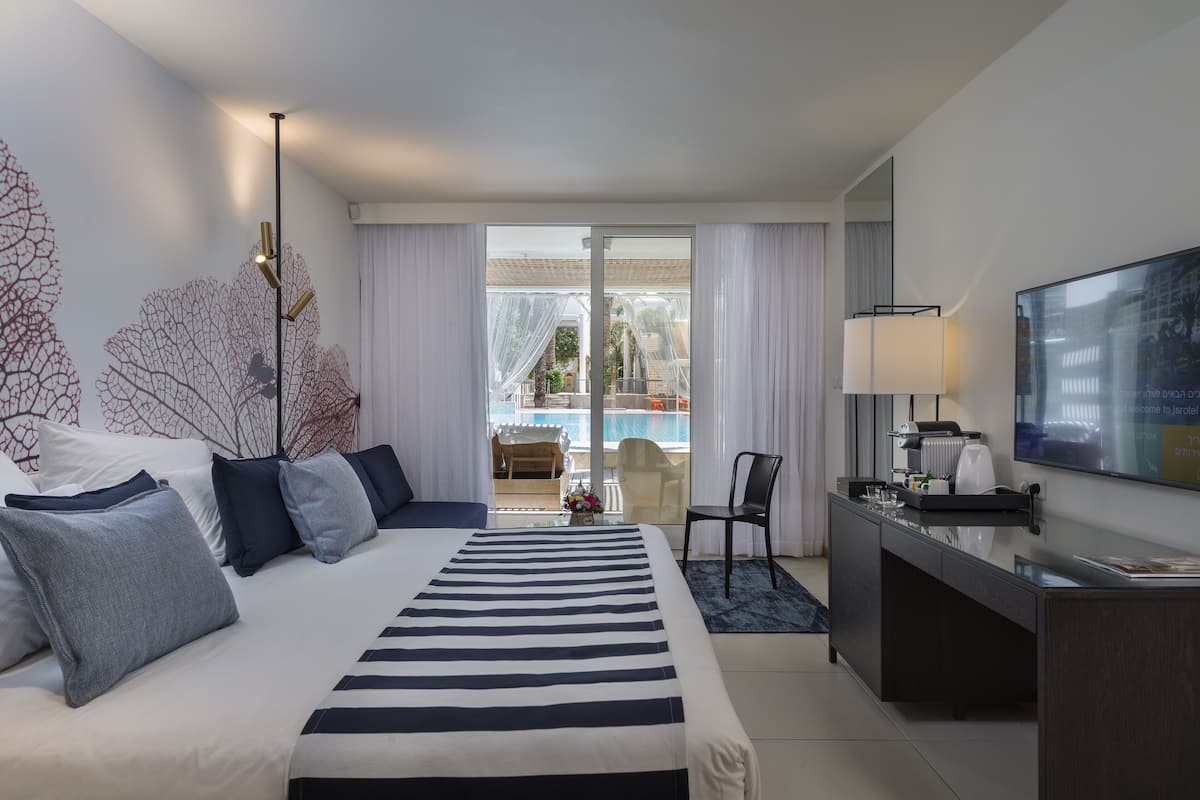 A hotel room with a large bed, a desk with a chair, a TV, and a view of a pool and outdoor area.